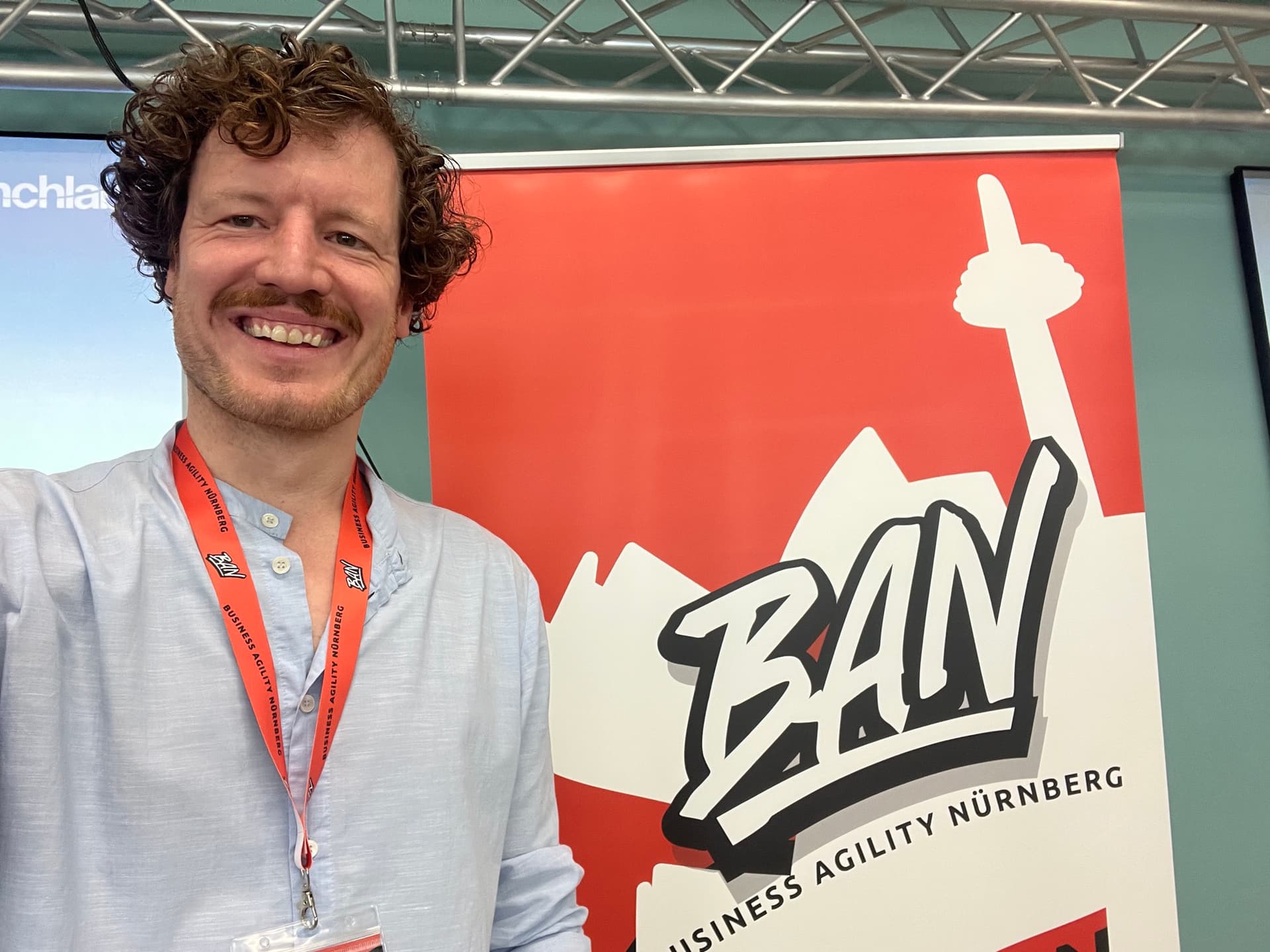 Dr. Simon Springmann lächelnd vor dem Roll-up der Business Agility Nürnberg mit BAN-Logo und Speaker-Lanyard