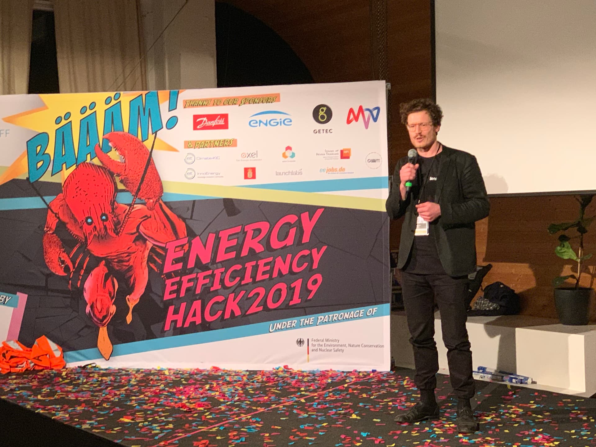 Dr. Simon Springmann als Jurymitglied auf der Bühne des DENEFF Energy Efficiency Hack 2019 in Berlin, vor Sponsorenwand mit Bundesumweltministerium-Patronage