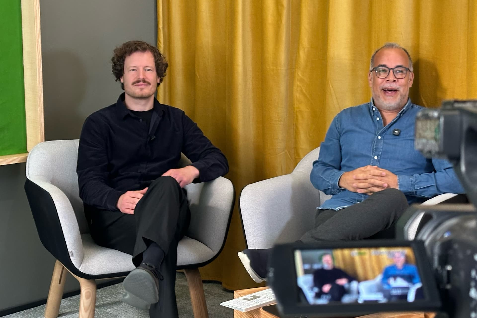 Dr. Simon Springmann im Sessel im launchlabs-Studio in Berlin, dahinter ein gelber Vorhang, im Vordergrund eine Filmkamera, beim Handelsblatt-Live-Seminar „KI x Innovation“ 2026