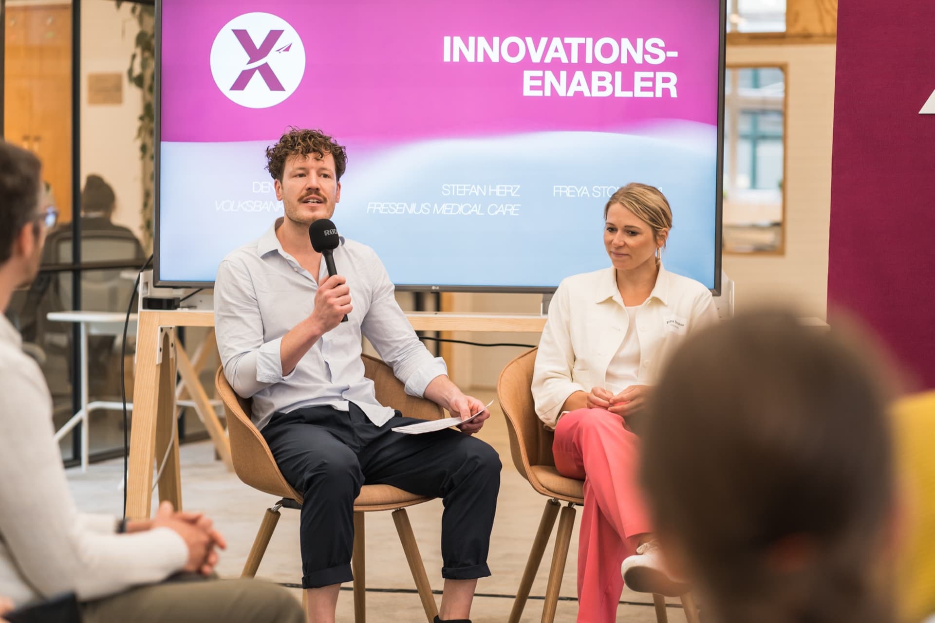 Dr. Simon Springmann moderiert das Innovations-Enabler-Panel beim LaunchX-Event 2023 in Berlin mit LAPP, Volksbank Lübeck und Fresenius Medical Care als Cases, sitzend mit Mikrofon vor pinkem Backdrop