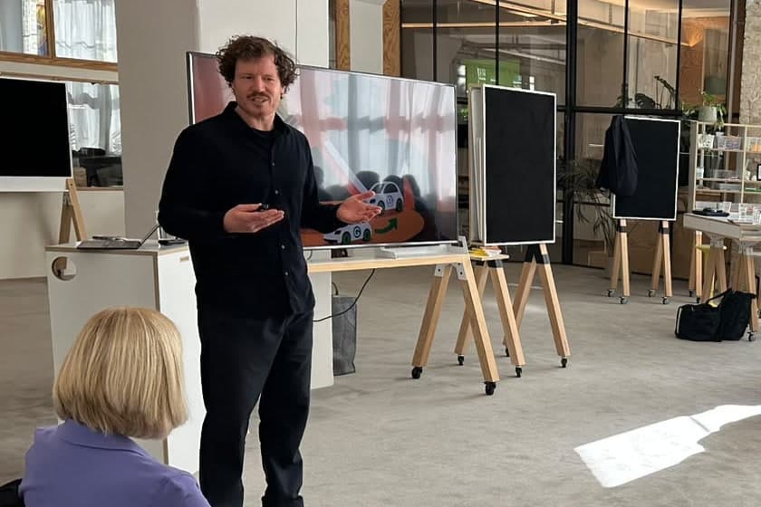 Dr. Simon Springmann gestikulierend vor einer Folie während seines Vortrags beim KI 360° in Berlin, im Vordergrund eine Zuhörerin von hinten