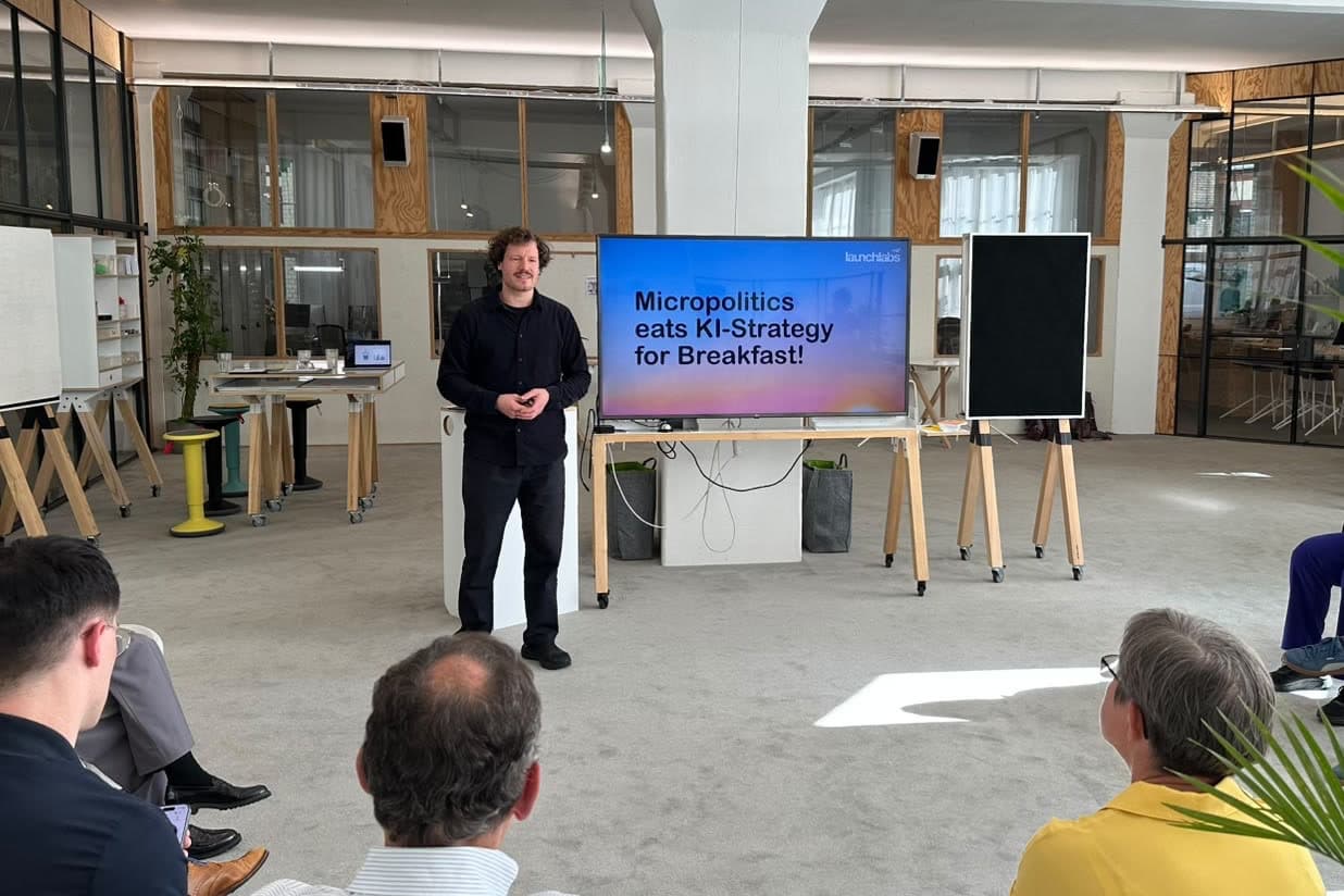 Dr. Simon Springmann neben seiner Titelfolie „Micropolitics eats KI-Strategy for Breakfast!“ beim KI 360° in den Räumen von launchlabs in Berlin, Publikum von hinten